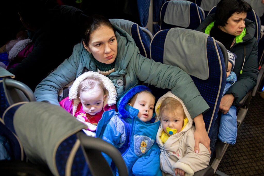 A woman sits with three small children in winter clothing on a coach, with other passengers nearby.