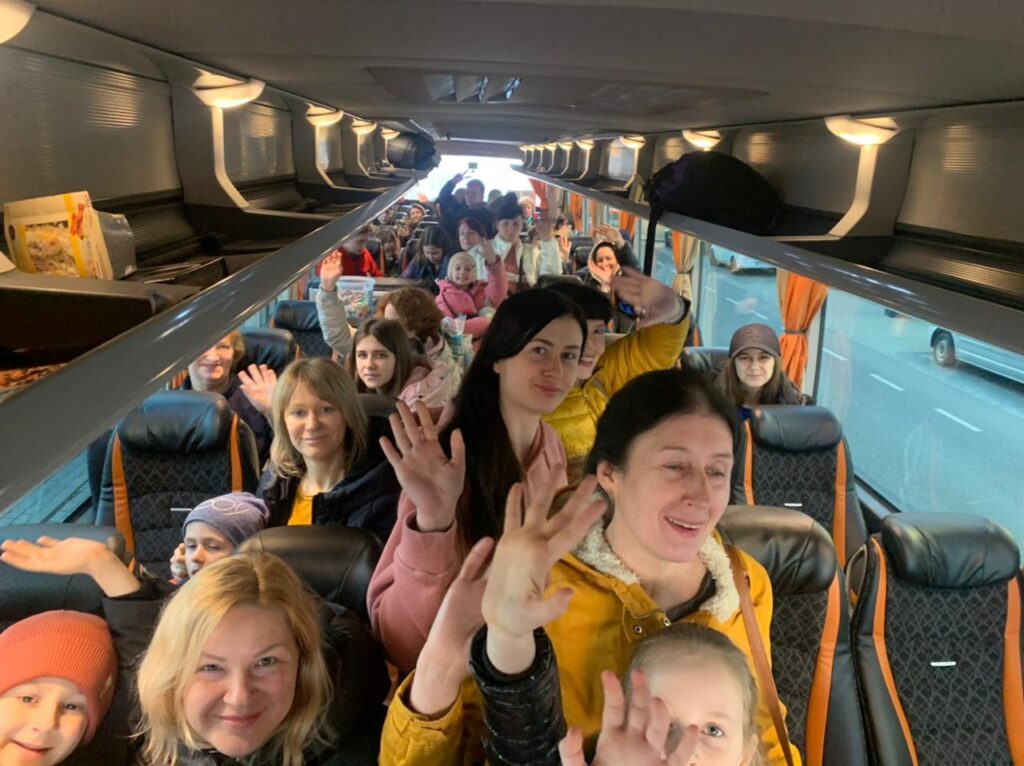 Women and children smile and wave from their seats on a full coach bus.