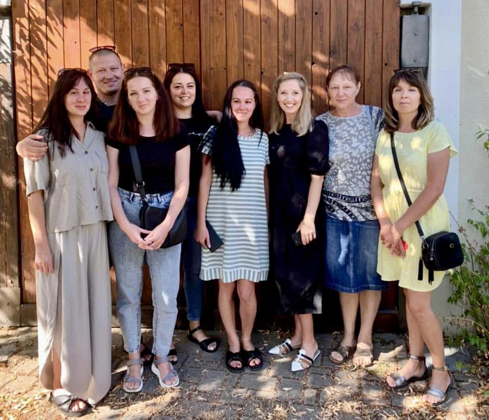 A group of eight people stand smiling together in front of a wooden door on a sunny day.